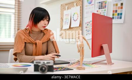 Asiatische Frau, die am Tisch sitzt und einen Stift hält und im Home Office auf den Computer schaut Stockfoto
