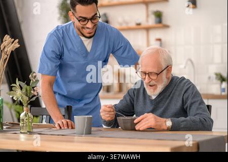 Lächelnder arabischer männlicher Pfleger mit Brille, der neben einem älteren Mann steht und beim Essen am Tisch zu Hause sitzt Stockfoto