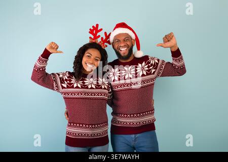 Fröhliches schwarzes Paar, das Weihnachtspullover trägt und Daumen auf sich selbst zeigt, während es im Studio vor blauem Hintergrund steht Stockfoto