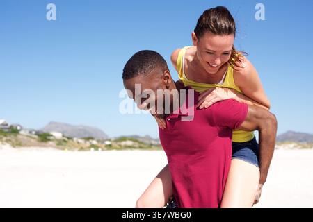 Fröhlicher schwarzer Mann, der an einem Sommertag am Strand zu einer kaukasischen Freundin mit Huckepack fährt Stockfoto
