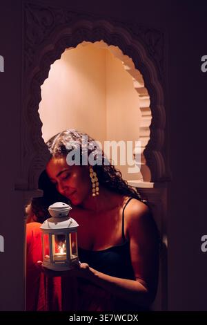 Arabische Frau, die Laterne mit brennender Kerze hält, sitzt nachts auf der Fensterbank des orientalischen Bogenfensters Stockfoto