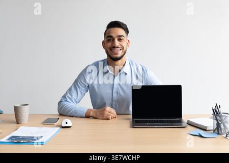 Ein lächelnder Mann aus dem Nahen Osten sitzt am Tisch mit Laptop im Büro Stockfoto