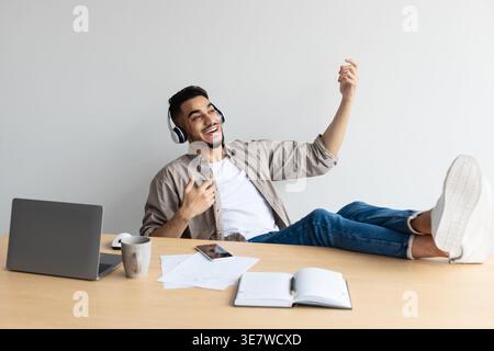 Mann aus dem Nahen Osten, der virtuelle Gitarre spielt und Musik mit Kopfhörern hört, sitzend mit den Füßen nach oben auf dem Tisch mit Laptop im Büro während der Pause Stockfoto