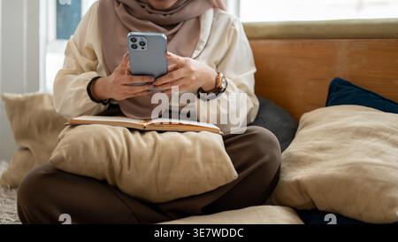 Asiatische muslimische Frau, die Hijab mit Smartphone trägt, während sie auf der Couch mit Kissen sitzt und im Wohnzimmer zu Hause Buch Stockfoto