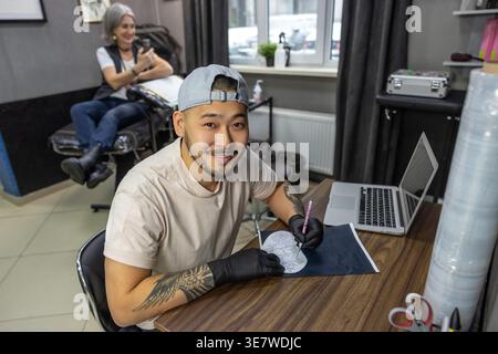 Lächelnder asiatischer Tätowierer mit Mütze und Latexhandschuhen, der an Skizzen arbeitet, während er in seinem Tattoo-Studio am Tisch sitzt Stockfoto