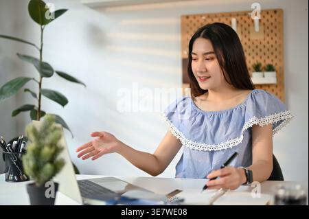 Lächelnde asiatische Unternehmerin, die mit einem Laptop an einem geschäftlichen Auftrag arbeitet, während sie im Heimbüro am Tisch sitzt Stockfoto