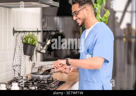 Lächelnder arabischer männlicher Pfleger in Brille kocht, während er zu Hause in der Küche steht Stockfoto