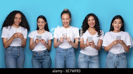 Verschiedene Frauengruppen, die weiße Hemden und Jeans tragen und Smartphones benutzen, die im Studio vor blauem Hintergrund stehen Stockfoto