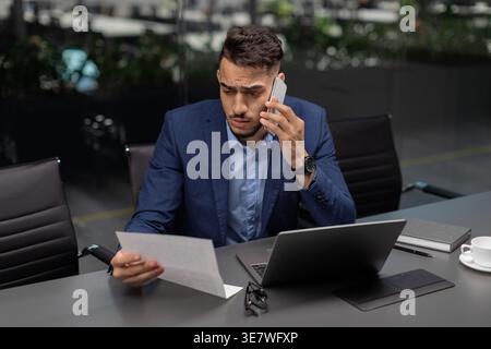 Ein gestresster arabischer Geschäftsmann liest Dokumente und telefoniert am Schreibtisch mit Laptop und Brille im Büro Stockfoto