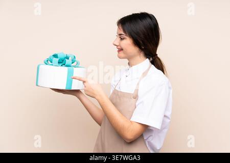 Seitenansicht einer hispanischen Patisserie, die in die Richtung zeigt und den Kuchen hält, der vor beigem Hintergrund im Studio steht Stockfoto