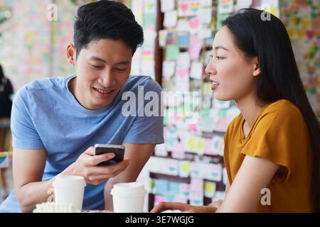 Asiatischer Mann, der mit seinem Smartphone sitzt, während er mit seiner Freundin im Café am Tisch sitzt Stockfoto