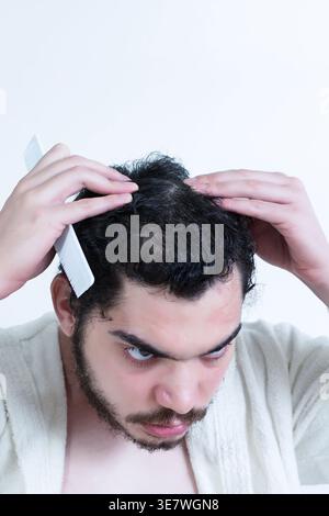 Mann aus dem Nahen Osten, der Bademantel trägt und die Haare überprüft, der Kamm hält, der zu Hause an der weißen Wand steht Stockfoto