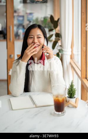 Thailändische Frau, die telefoniert und lachend wegschaut, während sie am Tisch mit einem Glas Eiskaffee im Café sitzt Stockfoto