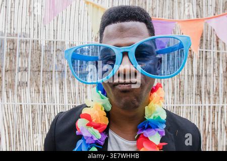 Schwarzer Mann mit großer Sonnenbrille und hawaiianischer Blumengirlande, die Gesicht vor der Wanddekoration während der Party im Freien macht Stockfoto