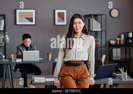 Lächelnde asiatische Geschäftsfrau lehnt sich an einen Tisch neben einem männlichen Kollegen im Büro Stockfoto