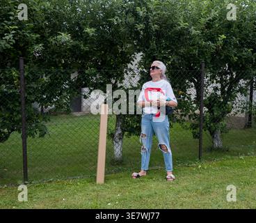 Eine ältere Frau in zerrissener Jeans steht an einem Zaun vor einem Obstgarten in Tschechien Stockfoto