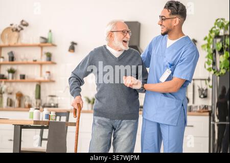 Lächelnder arabischer männlicher Betreuer in Brille, der einen älteren Mann im Stehen zu Hause ansieht Stockfoto