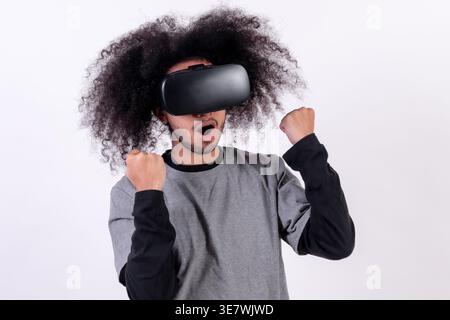 Hispanischer Mann mit lockigem Haar, der im Studio eine VR-Brille mit geballten Fäusten vor weißem Hintergrund trägt Stockfoto