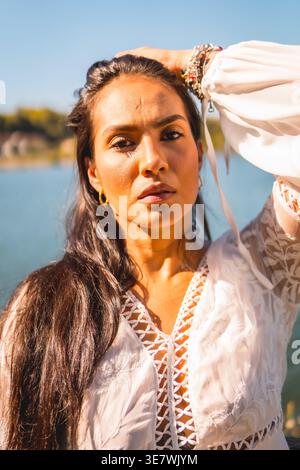 Ernsthafte hispanische Frau, die an sonnigem Tag mit der Hand auf dem Kopf gegen den See im Park steht Stockfoto