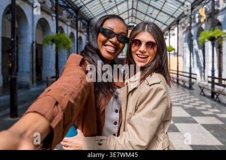 Schwarze und kaukasische Freundinnen tragen Trenchcoat und Sonnenbrille und machen Selfies, während sie tagsüber in der Stadt stehen Stockfoto