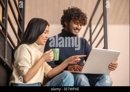 Hispanische Frau, die einen Becher hält und mit einem asiatischen Mann sitzt, der einen Laptop auf der Treppe benutzt Stockfoto