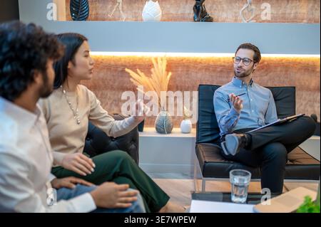 Asiatische weibliche und indische männliche Paare sitzen eng zusammen auf dem Sofa und unterhalten sich mit kaukasischen männlichen Psychotherapeuten in der Klinik Stockfoto