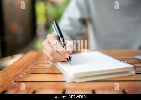 Zugeschnittenes Bild von thailändischen männlichen Schriften in Notizbuch, während sie an einem Holztisch in einem Café im Freien sitzen Stockfoto