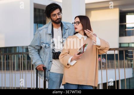 Asiatisches und kaukasisches Paar mit Brille, die Reisedokumente hält, während es dicht neben dem Gepäck vor dem Flughafen steht Stockfoto