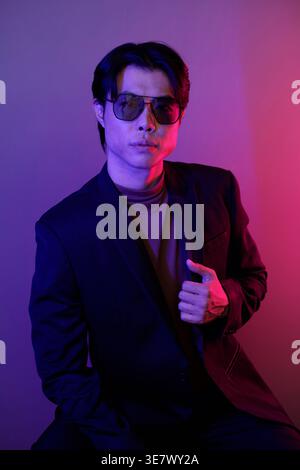 Asiatischer Mann mit Sonnenbrille, der auf Stuhl sitzt und seinen Mantel mit lila Lichteffekt auf dem Hintergrund im Studio hält Stockfoto