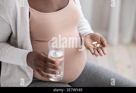 Schwarze schwangere Frau mit Vitaminpille Kapsel und Glas Wasser, während sie zu Hause auf dem Bett im Schlafzimmer sitzt Stockfoto
