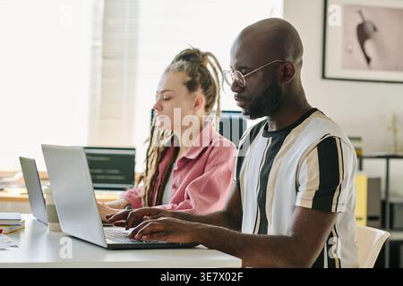 Schwarze männliche und kaukasische Programmierer geben Codes auf Laptops ein, während sie zusammen am Tisch im Büro sitzen Stockfoto