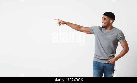 Fröhlicher Schwarzer Mann, der ein graues Hemd trägt, der wegzeigt, während er vor weißem Hintergrund im Studio steht Stockfoto