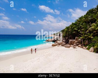 Ein Paar genießt einen romantischen Spaziergang entlang der atemberaubenden Sandküste von Anse Source d'Argent auf den Äußeren Inseln der Seychellen. Üppiges Grün und türkisfarbenes Wasser schaffen eine paradiesische Umgebung. Stockfoto