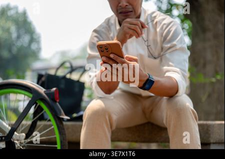 Trauriger asiatischer Geschäftsmann, der eine Brille mit einer Hand hält, indem er tagsüber auf einer Betonbank in der Nähe des Fahrrads im Park sitzt Stockfoto