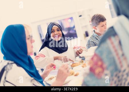 Lächelnde schwarze Frau im Hijab mit kaukasischen Freunden, die zusammen essen und zu Hause am Tisch sitzen Stockfoto
