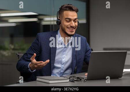 Arabischer Geschäftsmann, der ein Headset trägt, Videogespräche mit Kunden führt und ... auf den Laptop-Bildschirm blickt, während er mit Notebook und Brille am Schreibtisch sitzt Stockfoto