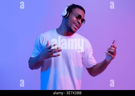 Sorgloser Schwarzer In Kabellosen Kopfhörern Und Sonnenbrillen Musik Auf Dem Smartphone Hören Und Singen, Lieblingslieder Genießen, Während Er In Neon Lig Steht Stockfoto