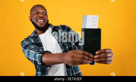 Fröhlicher schwarzer Mann mit Pass und Ticket, während er vor gelbem Hintergrund im Studio steht Stockfoto