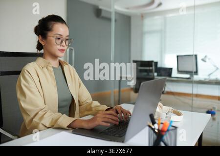 Asiatische Programmiererin, die eine Brille trägt und einen Laptop am Tisch im Büro benutzt Stockfoto