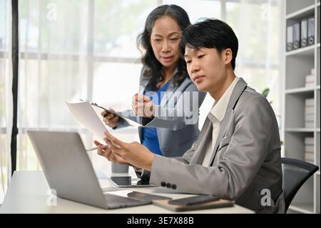 Asiatische Geschäftsfrau, die mit einem asiatischen Geschäftsmann an einem Finanzprojekt arbeitet, während sie im Büro auf einem Tisch sitzt Stockfoto