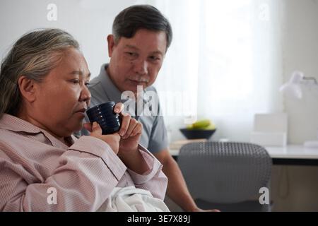 Seriöser älterer asiatischer Mann, der seine kranke Frau beim Trinken von grünem Tee ansieht, nachdem er Medizin genommen hat, während er zu Hause saß Stockfoto