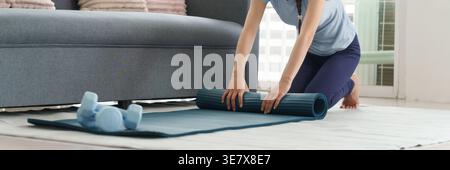 Erwachsene asiatische Frau rollt Yoga Matte Vorbereitung zu Hause Fitness Sitzung. Kurzhanteln in der Nähe zum Sport. Gesunde Lebensweise Praxis Selbstpflege. Stockfoto