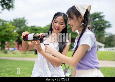 Thailändische Frau gießt Wein in ein Glas einer Freundin, die tagsüber auf einer Picknickdecke im Park sitzt Stockfoto