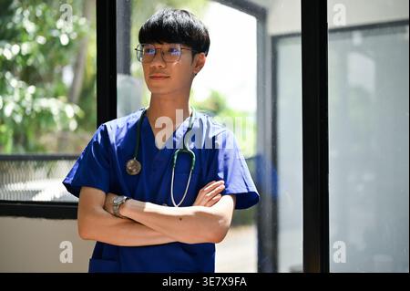 Seriöser asiatischer Arzt in blauen Peelings mit Stethoskop wegschauend mit Armen im Krankenhauskorridor überkreuzt Stockfoto