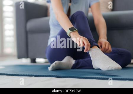 Erwachsene asiatische Frau trägt Aktivewear-verstellbare graue Socke auf einer blauen Trainingsmatte in ihrem modernen Wohnzimmer, die sich auf das Training vorbereitet. Stockfoto