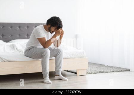 Gestresster bärtiger indischer Mann mit lockigem Haar, der die Stirn auf geballten Fäusten lehnt und im Schlafzimmer zu Hause auf dem Bett sitzt Stockfoto