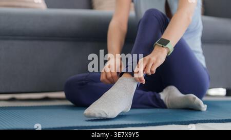 Asiatische Frau passt die graue Socke auf der Yogamatte an. Mit Sportswear-Smartwatch. Vorbereitung auf Fitness oder Entspannung in gemütlicher Wohnumgebung. Stockfoto