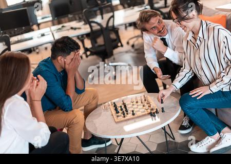 Enttäuschter Mann aus dem Nahen Osten, der das Spiel verlor, das sein Gesicht mit Händen bedeckt und Schach spielt, mit kaukasischen Arbeitskollegen, die im Büro auf dem Stuhl sitzen... Stockfoto