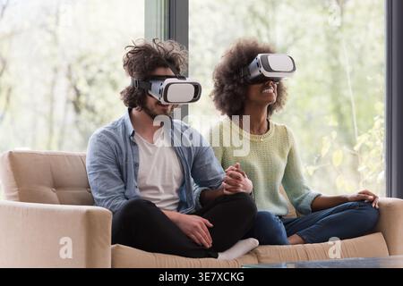 Schwarzes und kaukasisches Paar mit VR-Headsets, die Hände halten, während sie zusammen auf dem Sofa sitzen und virtuelle Realität im Wohnzimmer mit gl erleben... Stockfoto