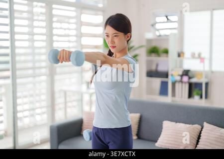 Erwachsene asiatische Frau, die in Fitnesstraining mit leichten Kurzhanteln in Innenräumen für persönliches Wohlbefinden und Kraftaufbau engagiert ist Stockfoto